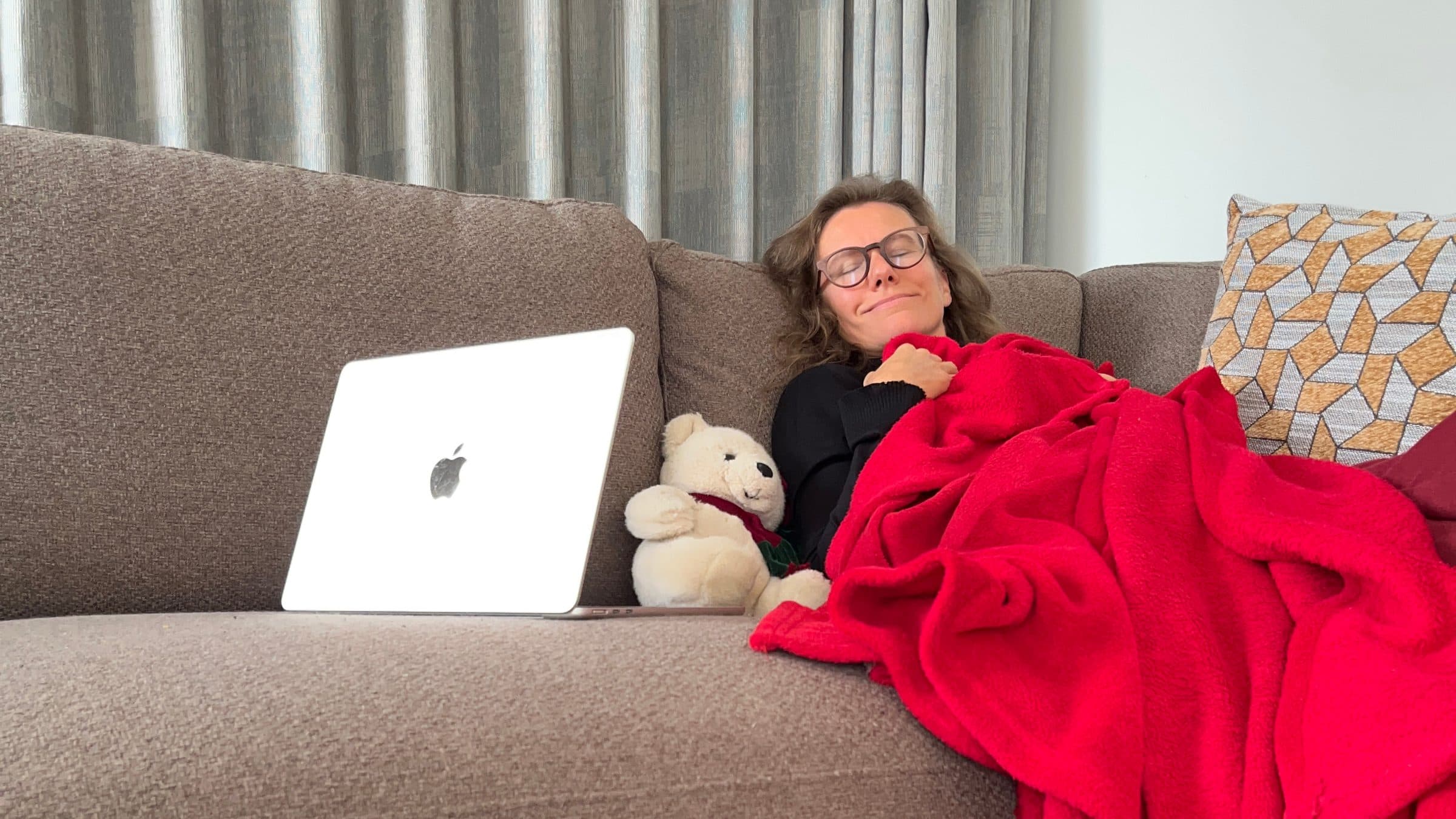 Elke Aerts ligt ontspannen met de ogen dicht onder een rood dekentje in de zetel, met haar laptop naast zich