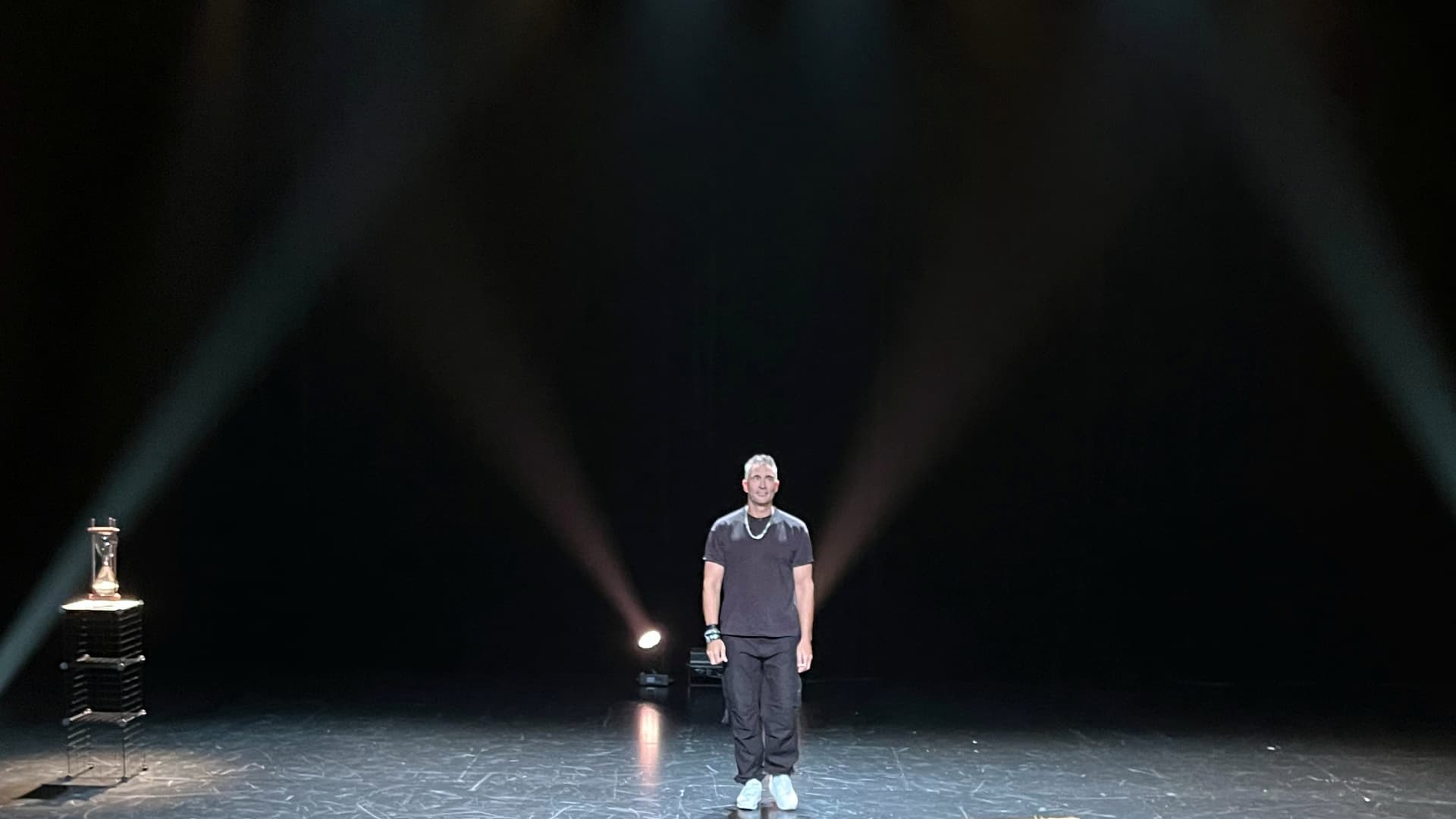 Comedian Guido Weijers alleen op een donker podium in het spotlicht, met links een zandloper op een sokkel